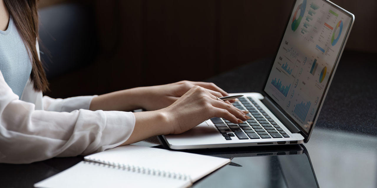 A professional Asian woman types on a laptop, analyzing data and taking notes, showcasing productivity and focus in a contemporary workspace.